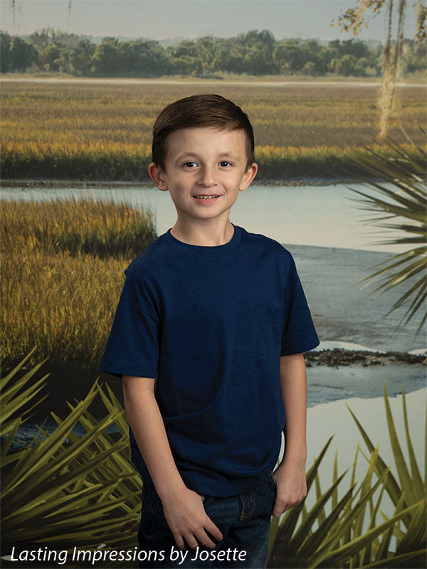 Coastal Marsh View photography backdrop showing golden marsh grass and a tidal creek with a distant treeline and palmetto fronds, shown with a young boy in a navy t-shirt standing and smiling CP-8486