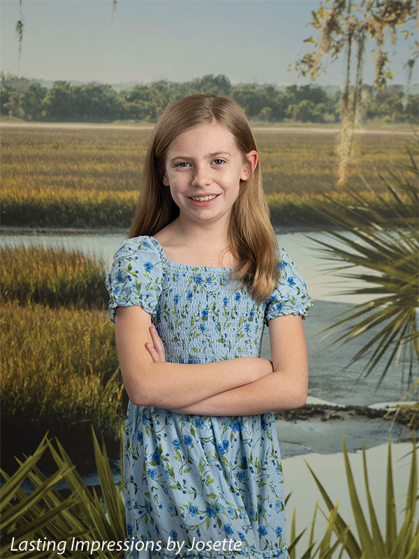 Coastal Marsh View photography backdrop showing golden marsh grass and a tidal creek with a distant treeline and palmetto fronds, shown with a young girl in a blue floral dress with her arms crossed CP-8486