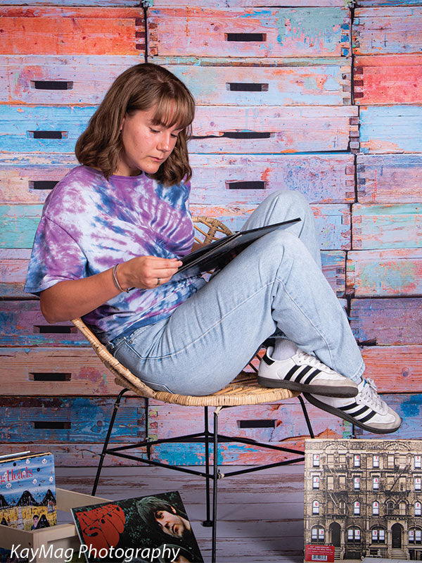 Teen girl seated in a rattan chair reading an album against a vibrant, painted wooden crate backdrop, perfect for retro or lifestyle-themed senior portraits.