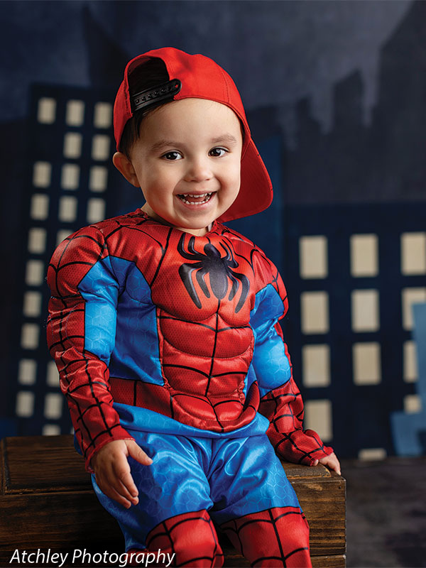 A toddler wearing a red and blue superhero costume and a red cap sits on a wooden box smiling, with a comic-style nighttime city backdrop featuring glowing windows behind him.
