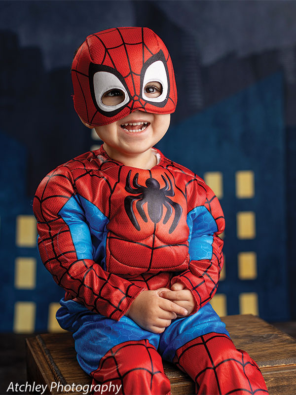 A smiling toddler wearing a red and blue superhero costume with a matching mask sits on a wooden box, hands folded, in front of a stylized night city backdrop with illuminated building windows.
