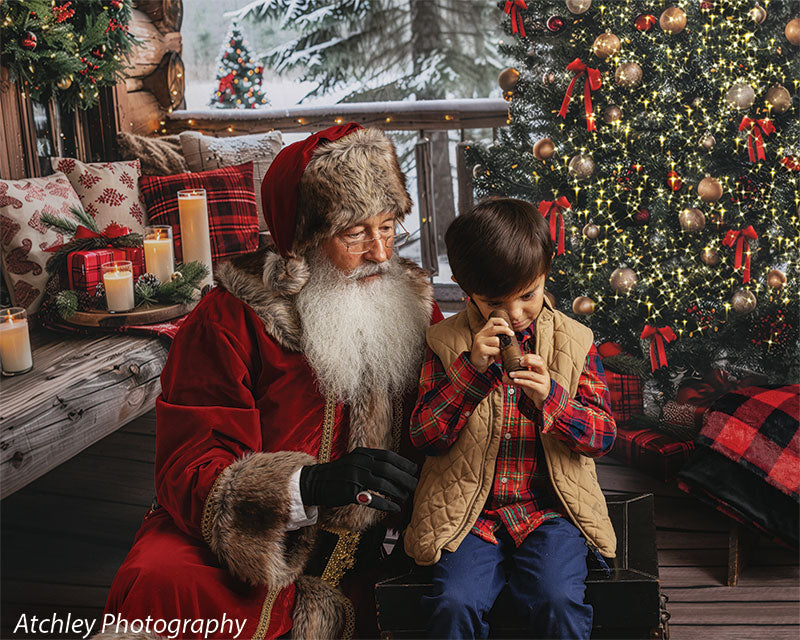 Santa Claus in a red fur trimmed suit sitting beside a young boy wearing a red plaid shirt, tan quilted vest, and blue pants on a wooden porch, with a decorated Christmas tree, glowing lights, wreath, candles, and snowy pine trees in a cozy log cabin Christmas backdrop.