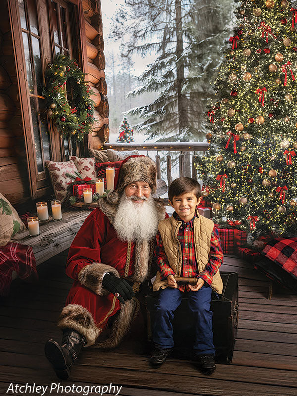 Santa Claus in a red suit with fur trim and a long white beard sitting beside a young boy wearing a red plaid shirt, tan quilted vest, and blue pants on a wooden porch, with a decorated Christmas tree, glowing lights, wreath on a log cabin wall, candles, and snowy trees in the cozy log cabin Christmas backdrop.