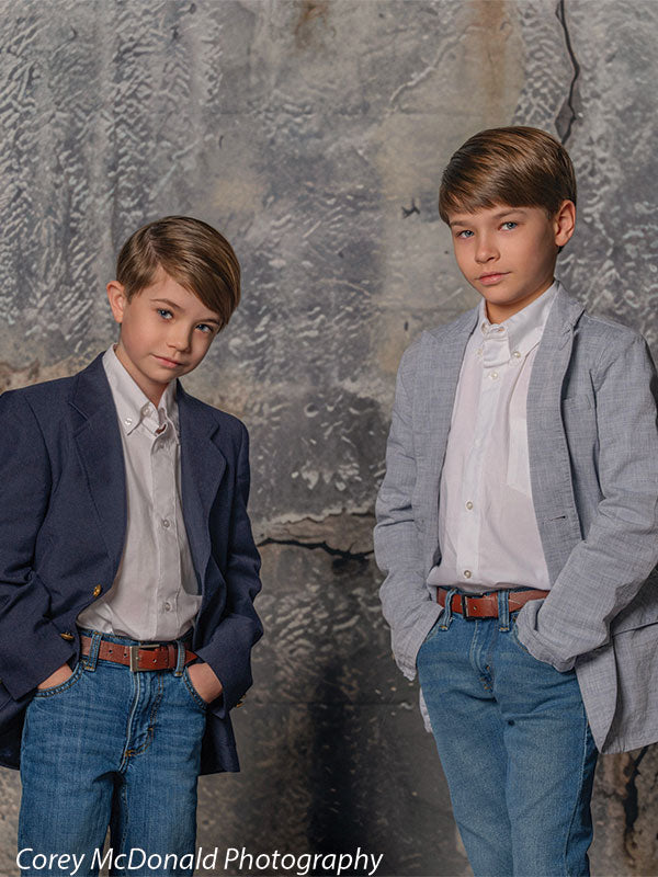 Two young boys wearing button-down shirts, blazers, and jeans stand side by side with hands in pockets in front of a cracked gray concrete wall backdrop with urban textured detail.
