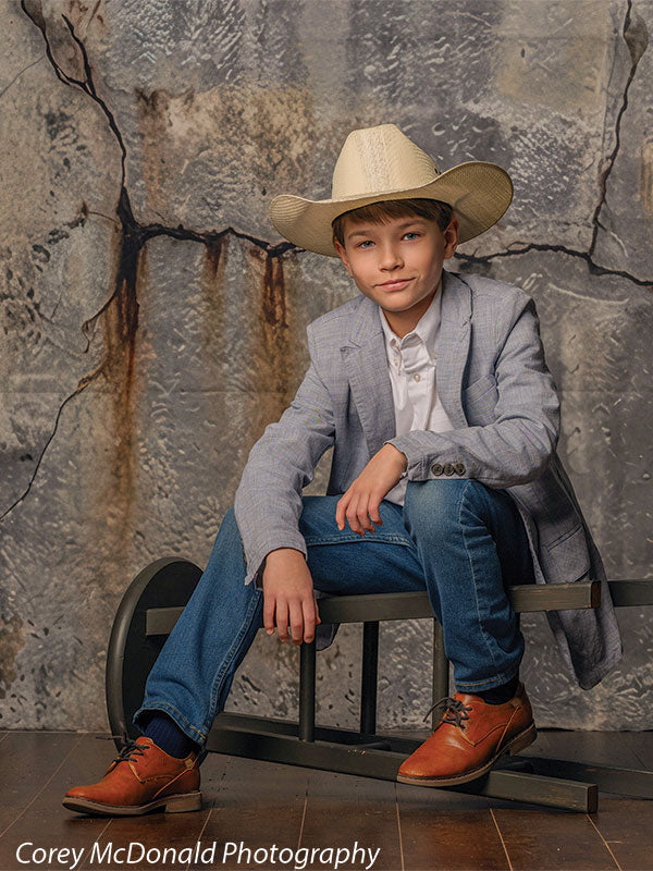 Young boy wearing a light gray blazer, white shirt, blue jeans, brown shoes, and a tan cowboy hat sits on a low bench with one arm resting on his knee in front of a cracked gray concrete wall backdrop with weathered texture.