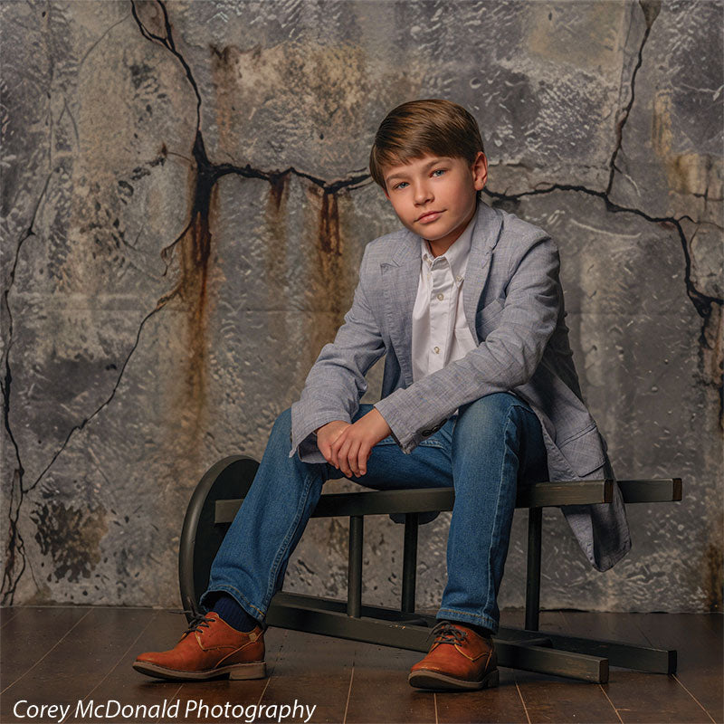 Young boy wearing a light gray blazer, white shirt, and jeans sits on a bench in front of a cracked industrial concrete wall backdrop with textured gray surface and visible fissures.
