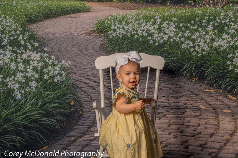 Baby girl in a yellow dress with blue detailing and a white bow headband sits on a white wooden chair placed on a curved brick garden path backdrop surrounded by blooming white flowers and lush greenery.
