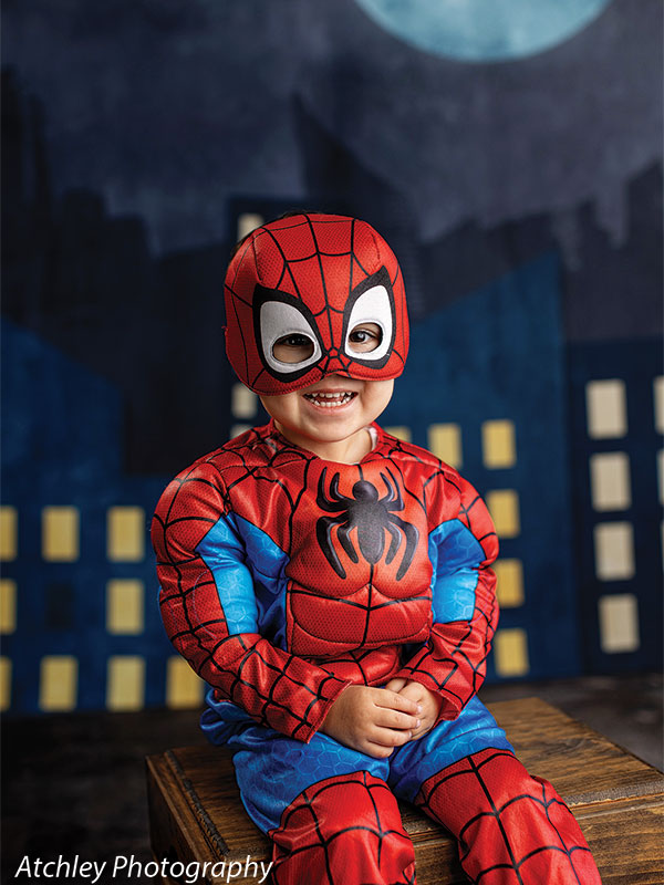 A toddler in a red and blue superhero costume with a mask sits on a wooden box, smiling toward the camera, posed against a dark city building backdrop under moonlight.
