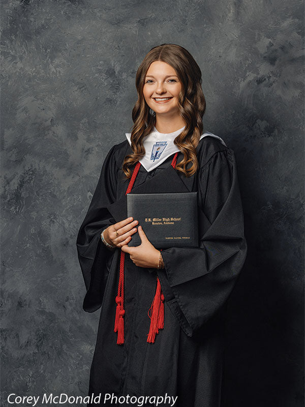 Teen girl with long wavy light brown hair wearing a black graduation gown with red honor cords smiles while holding a diploma in front of a dark gray mottled photography backdrop with textured effect.