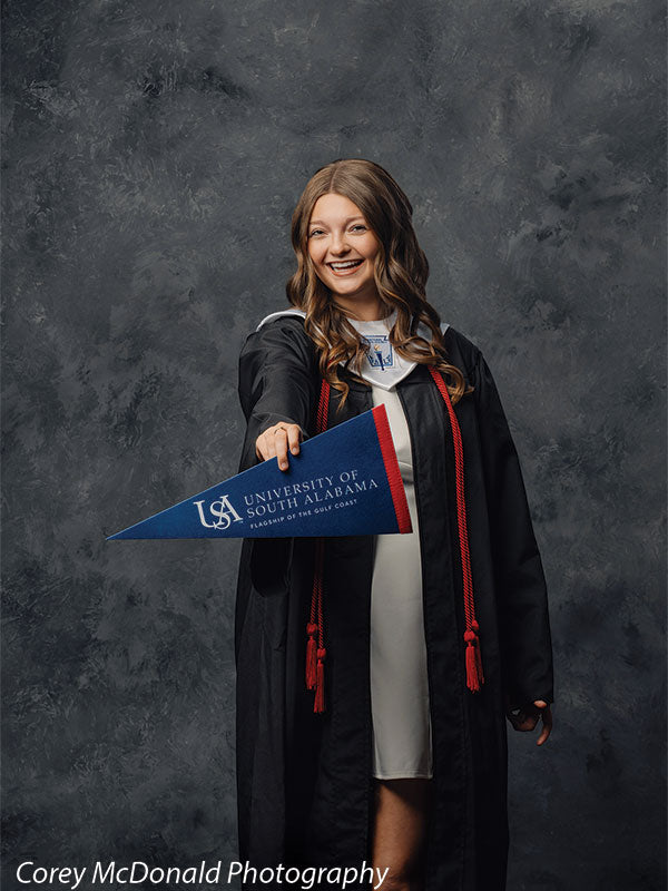 Teen girl with long wavy light brown hair wearing a black graduation gown with red cords holds a blue University of South Alabama pennant extended forward with a wide smile in front of a dark gray painted studio backdrop.