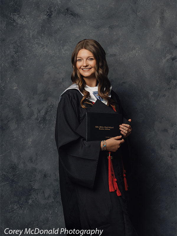 Teen girl with long wavy light brown hair wearing a black graduation gown with red honor cords stands in an angled pose holding a diploma and smiling in front of a dark gray studio backdrop with a subtle mottled pattern.