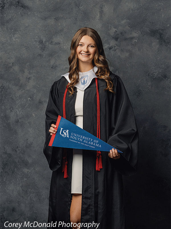 Teen girl with long wavy light brown hair wearing a black graduation gown with red cords holds a blue University of South Alabama pennant while standing centered in front of a dark gray textured backdrop.
