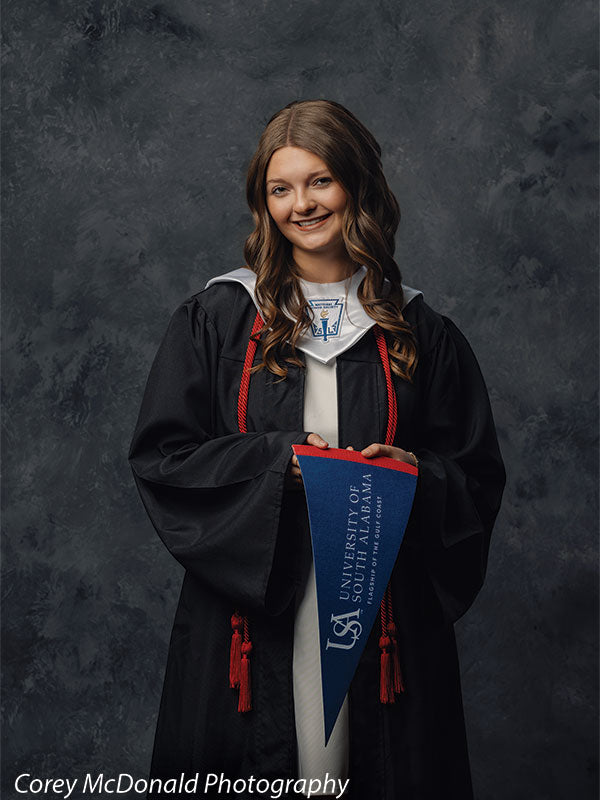 Teen girl with long wavy light brown hair wearing a black graduation gown with red cords holds a blue University of South Alabama pennant downward while smiling in front of a dark gray textured photography backdrop with soft tonal variation.