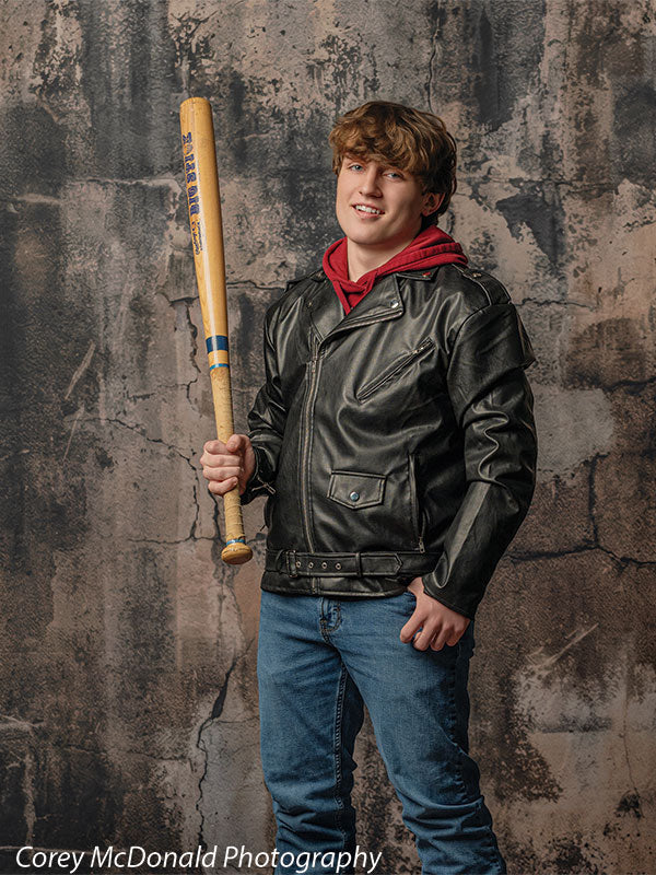 Teen boy dressed in a black leather jacket, red hoodie, and blue jeans holds a wooden baseball bat upright while smiling in front of a dark grunge concrete wall backdrop with rough industrial texture.