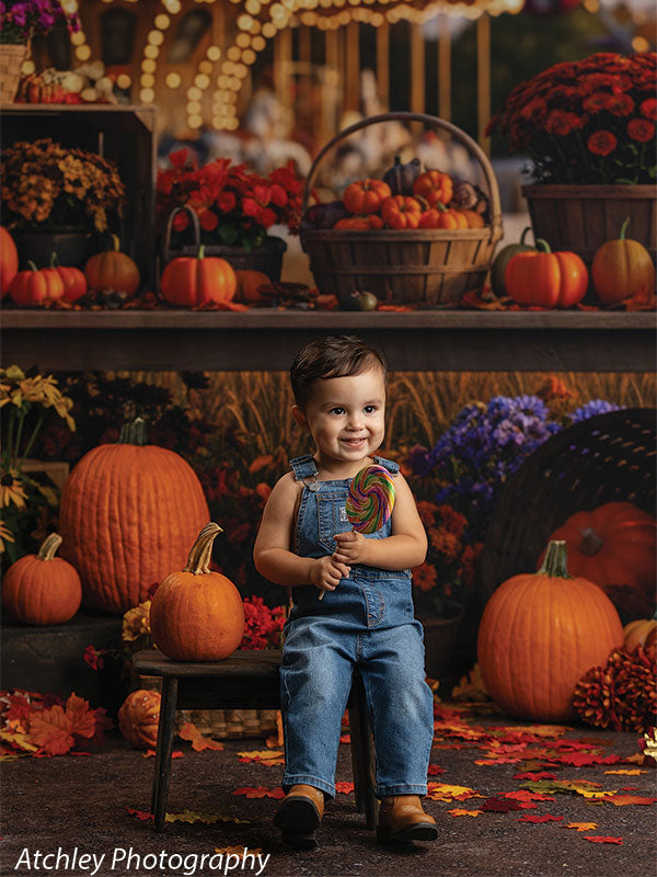Smiling toddler in blue denim overalls seated on a dark stained rustic wooden kids bench holding a rainbow swirl lollipop, posed among large pumpkins and fallen leaves on a dirt look floordrop, with a nostalgic carnival fairground backdrop filled with harvest baskets, flowers, and glowing carousel lights.