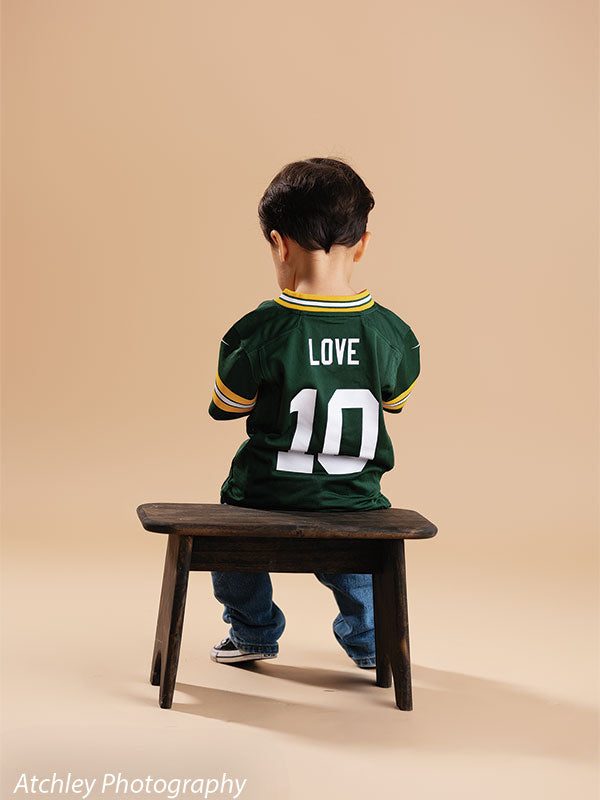 Toddler sitting on a dark stained wooden kids bench facing away, wearing a green football jersey with Love and number 10 on the back, blue jeans, and sneakers, against a khaki brown seamless background.