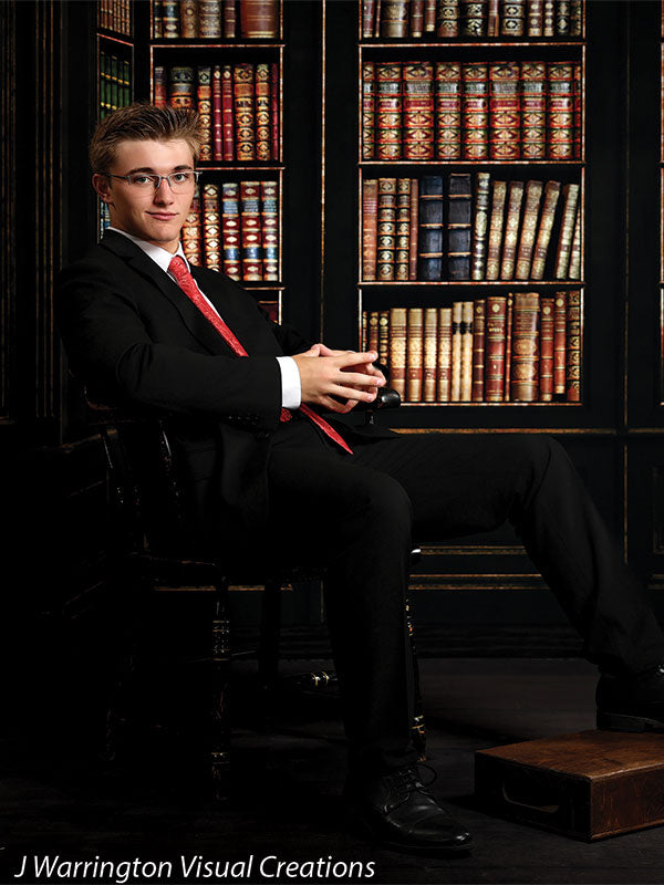 Teen boy in formal black suit seated on a vintage chair over a dark wood floordrop, styled for academic portraits with classic flooring aesthetics.