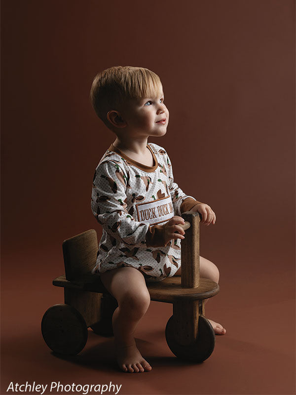 A toddler wearing a patterned romper sits barefoot on a wooden scooter toy, looking upward while holding the handle, posed against a deep brown minimal studio backdrop.
