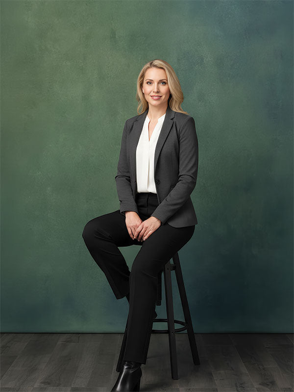 A woman wearing a dark blazer and white blouse sits on a stool with one leg crossed, posed against a deep green textured studio backdrop with subtle tonal variation and a professional portrait style.
