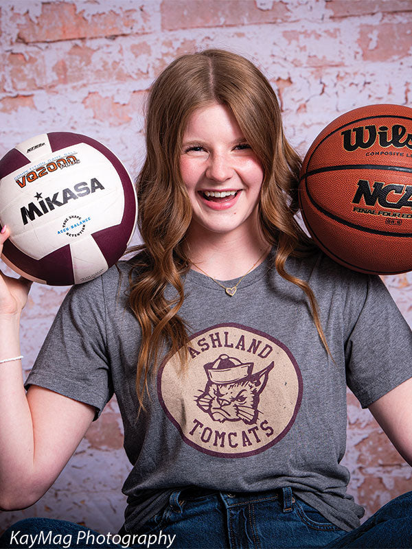 Close-up of a smiling teen holding volleyball and basketball in front of a pink-toned distressed brick backdrop, styled for team or school spirit photography.