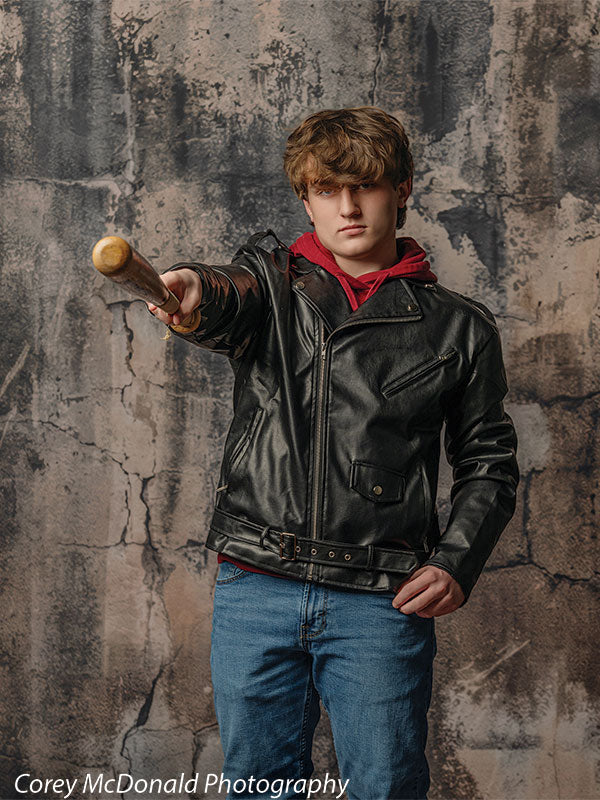 Teen boy wearing a black leather jacket and red hoodie points a wooden baseball bat forward while standing against a cracked gray concrete wall backdrop with a distressed industrial look.