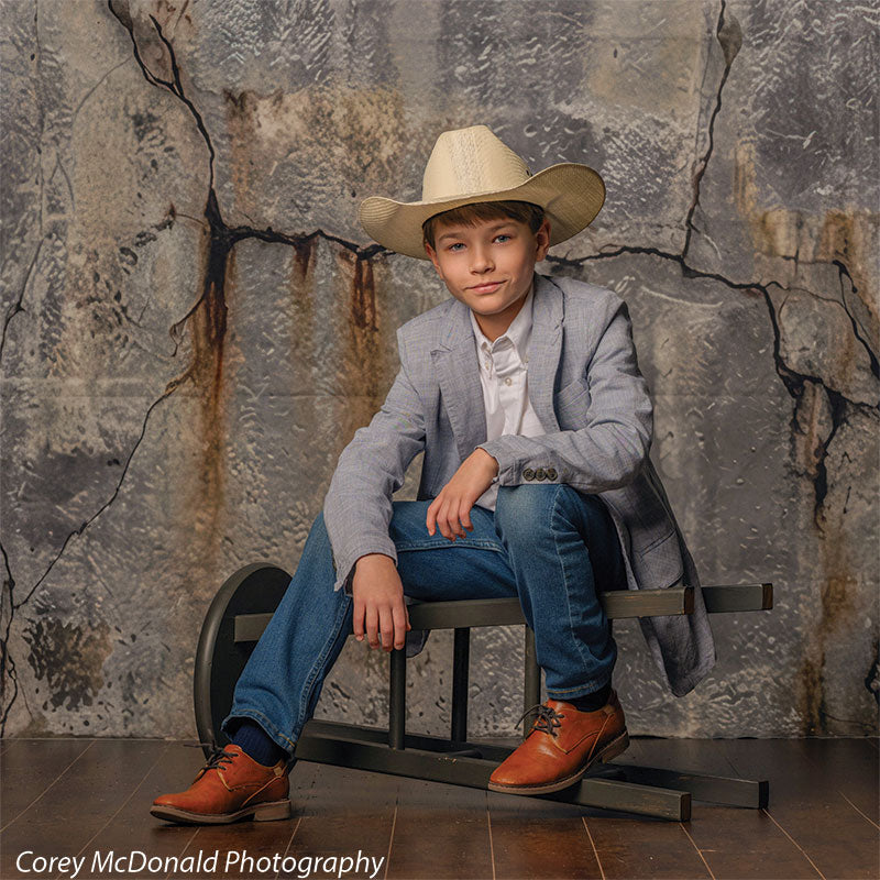 Young boy in a light gray blazer, white shirt, blue jeans, brown shoes, and a tan cowboy hat sits casually on a bench with legs apart in front of a distressed gray concrete wall backdrop featuring visible cracks and industrial texture.