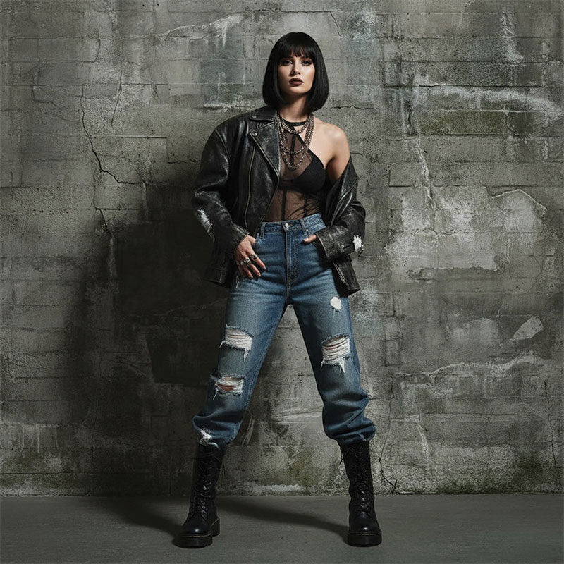 Woman with black bob hairstyle in a leather jacket, sheer top, distressed blue jeans, and boots poses confidently with hands near pockets in front of a distressed gray urban concrete wall backdrop.