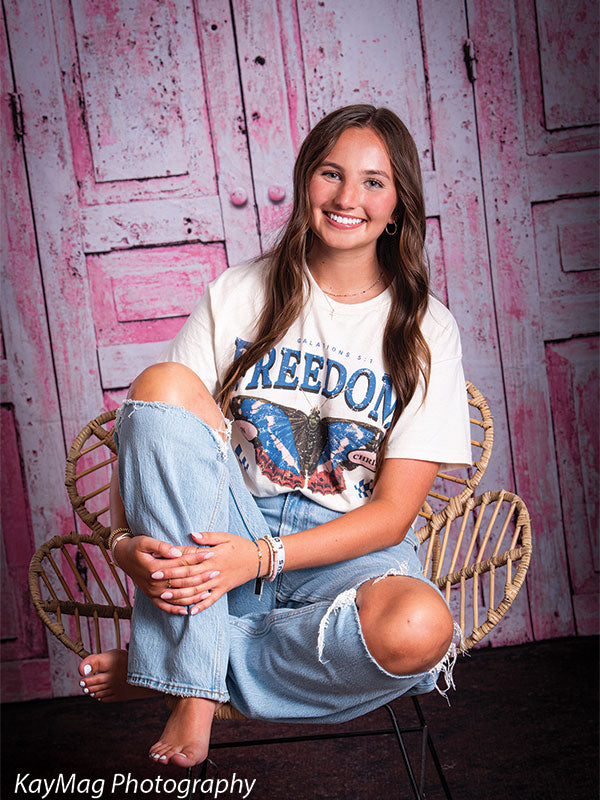 Barefoot teen girl sitting cross-legged on a wicker chair with dark flooring beneath a distressed pink wood backdrop, great for casual or boho-themed portraits.