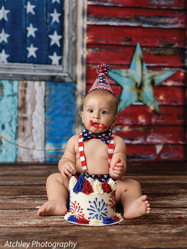 Baby wearing a patriotic cone hat with pom pom, star bow tie, and red striped suspenders sitting on a wooden floor with red frosting on their mouth, in front of a distressed red white and blue wooden American flag backdrop with a large teal star, holding a decorated red white and blue cake.