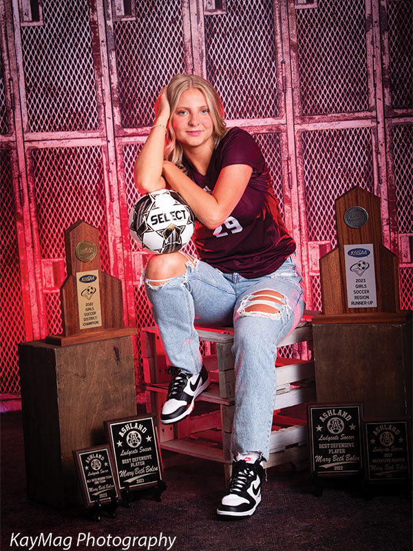 Rustic wood apple boxes used as props in a sports-themed senior portrait featuring a soccer player with trophies against a red metal locker backdrop.