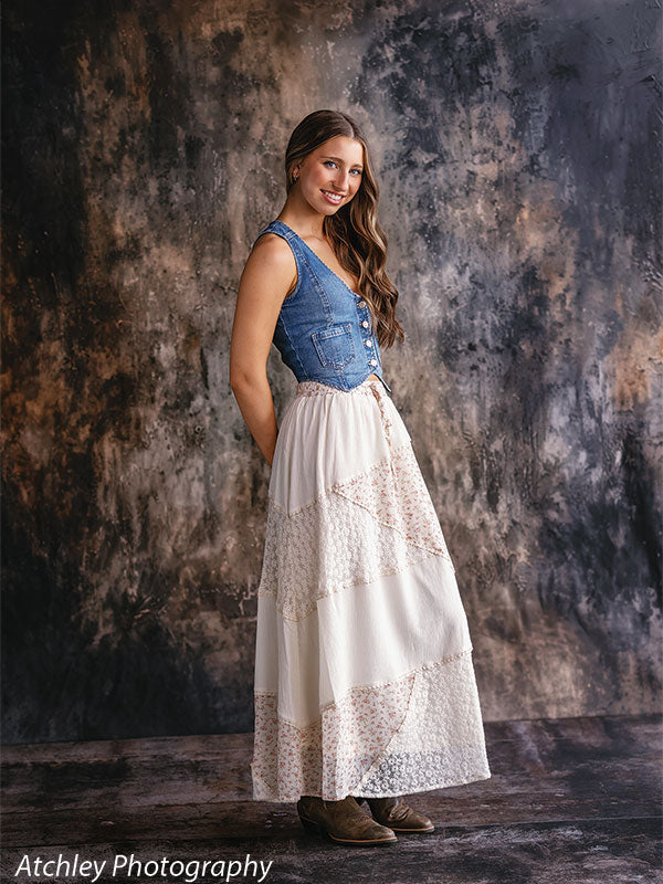 Young woman standing in profile wearing a denim vest, white lace maxi skirt, and brown boots, posed against an earth tone abstract portrait photography backdrop with blended gray and brown textures.