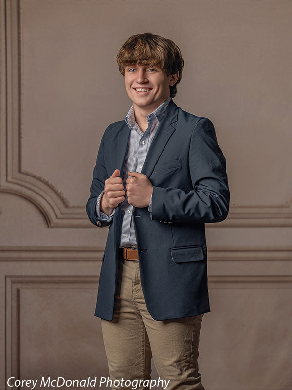 Teen boy with wavy light brown hair wearing a blue blazer, light blue shirt, and khaki pants smiles while adjusting his jacket, standing in front of an elegant beige wall panel backdrop with classic portrait styling.