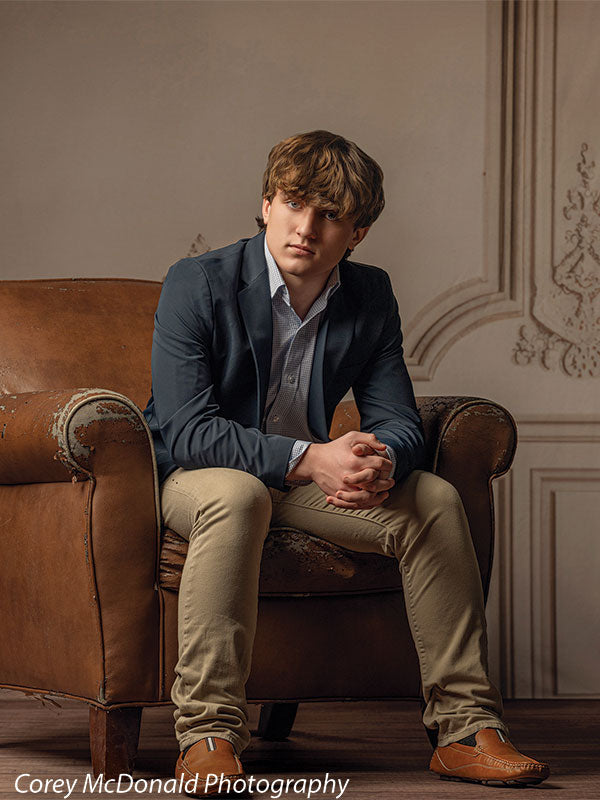 Teen boy in a navy blazer, light blue shirt, khaki pants, and brown shoes sits forward in a worn brown leather armchair with hands clasped in front of a beige vintage wall panel backdrop with ornate molding details.