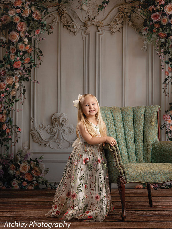 A young girl in a floral embroidered dress stands beside a green upholstered chair, smiling, with an elegant floral wall backdrop and wooden floor beneath her.
