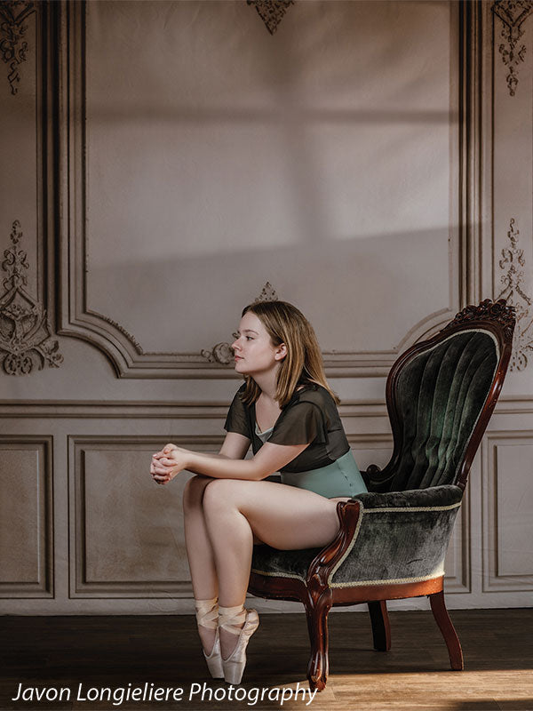Elegant French Panel Wall photography backdrop with an antique green velvet armchair, shown with a young ballet dancer seated on the chair with hands clasped in a wide studio view CP-8744