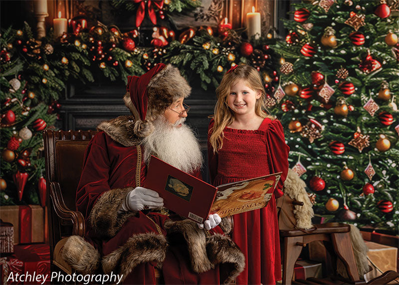 Santa Claus reading from a large Christmas book while a smiling young girl in a red dress stands beside him, posed in front of an elegant holiday fireplace backdrop with a decorated tree, wreath, garland, candles, and wrapped presents.