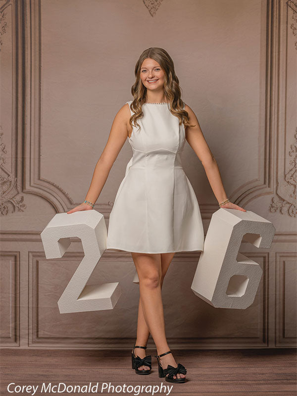Teen girl with long wavy light brown hair wearing a sleeveless white dress stands smiling while holding large white numbers two and six in front of an elegant neutral wall backdrop with classic panel molding.