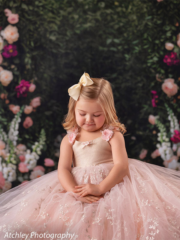 A young girl wearing a blush pink tulle dress looks down with hands folded in her lap, posed in front of a dark green rose garden backdrop with soft floral accents.
