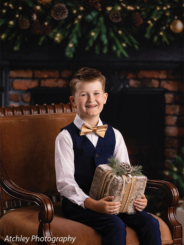Ember & Pine brick fireplace with Christmas garland photography backdrop shown with a boy in a navy vest and gold bow tie seated holding a wrapped gift CP-8881