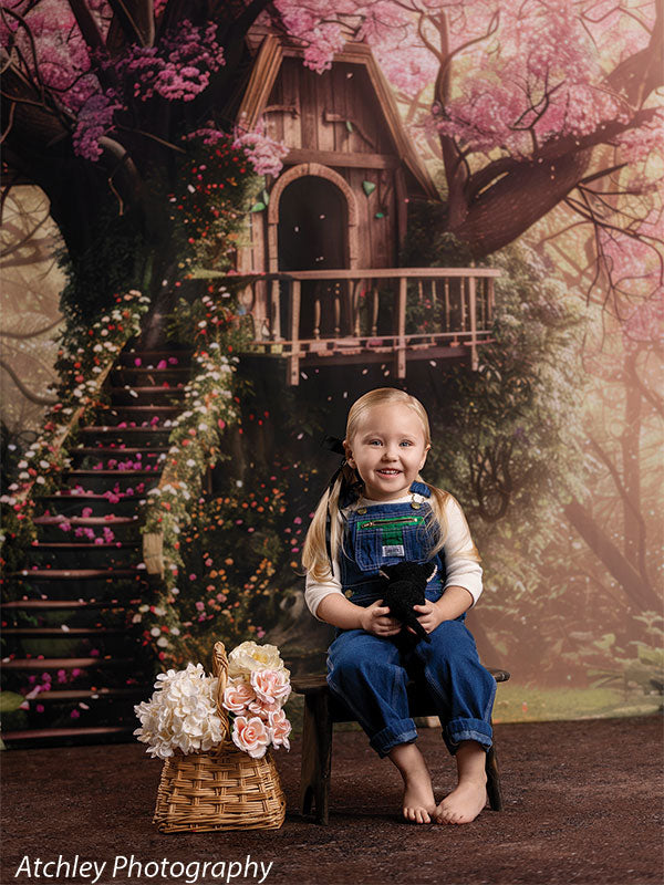 A young girl wearing denim overalls and a white shirt sits barefoot on a small chair holding a stuffed animal, posed in front of an enchanted forest treehouse backdrop with pink blossoms and winding stairs.
