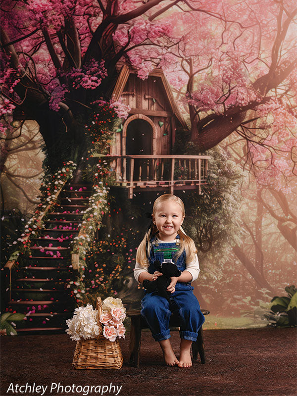 A young girl wearing denim overalls and a white shirt sits barefoot on a small chair holding a stuffed animal, with a basket of flowers beside her, in front of an enchanted forest treehouse backdrop with pink blossoms.
