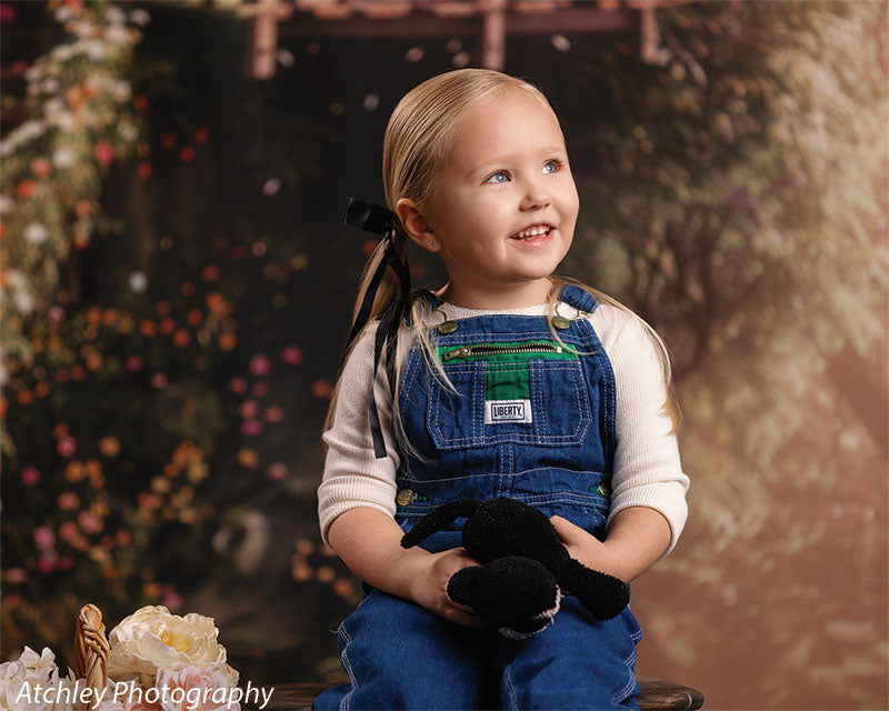 A young girl in denim overalls and a white shirt sits holding a stuffed animal and smiling while looking upward, posed in front of a fairytale-style spring forest treehouse backdrop with flowers and foliage.