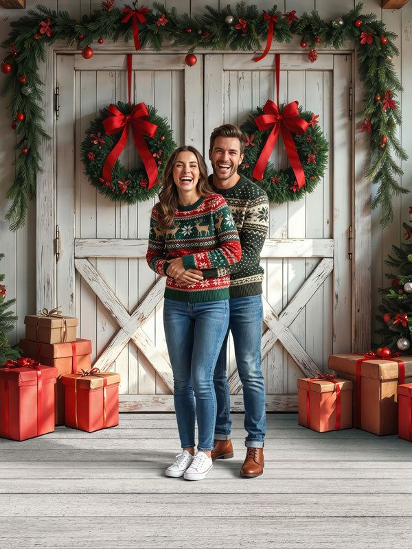 Farmhouse Noel white barn doors with paired Christmas wreaths photography backdrop shown with a couple in matching sweaters standing together CP-8933