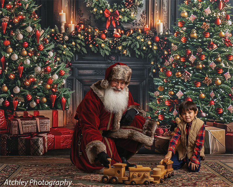Santa Claus kneeling beside a young boy playing with wooden toy trains on a patterned rug, surrounded by wrapped presents and a decorated Christmas tree, with a festive holiday fireplace backdrop featuring garland, wreath, candles, and glowing lights.