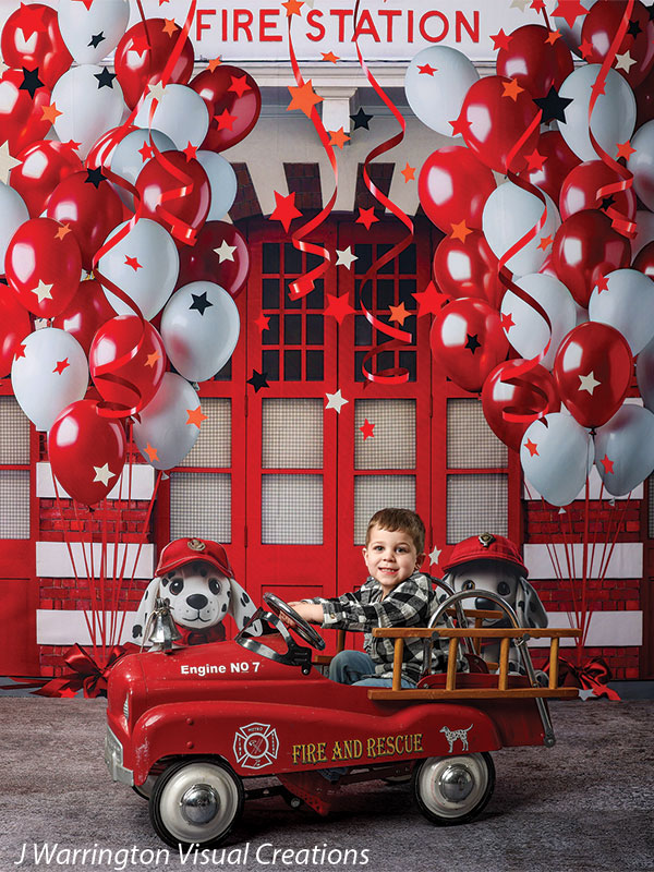 Young boy riding a red firetruck pedal car in front of a festive firehouse cake smash backdrop with red and white balloons and firefighter dalmatian plush toys.