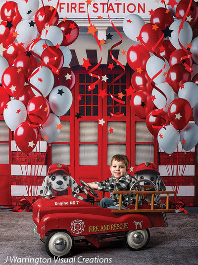 Young boy riding a red firetruck pedal car in front of a festive firehouse cake smash backdrop with red and white balloons and firefighter dalmatian plush toys.