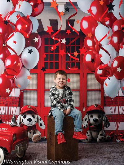 Smiling boy sitting on a wooden stool in front of a bright red firehouse backdrop decorated with red and white balloons and firehouse plush toys.