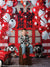 Smiling boy sitting on a wooden stool in front of a bright red firehouse backdrop decorated with red and white balloons and firehouse plush toys.