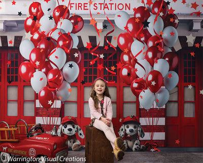 Smiling girl posing in front of a full view of the firehouse cake smash backdrop featuring festive balloons, red garage doors, and dalmatian plush toys.