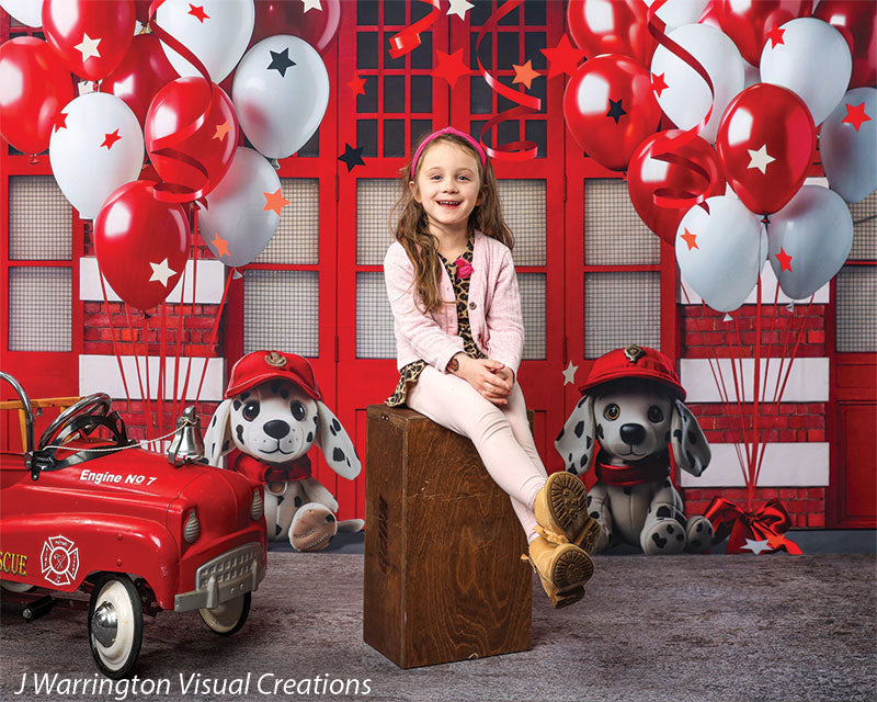 Girl sitting on a stool in front of a firehouse-themed backdrop with red and white balloons and firefighter stuffed animals.